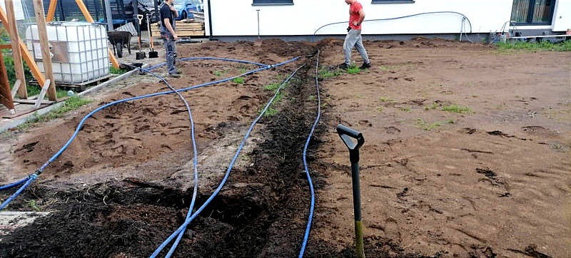 Wykonanie drenażu w ogrodzie: wykopany rów, niebieskie rury drenażowe ułożone wzdłuż rowu, pracownicy w tle, przygotowanie podłoża pod trawnik.