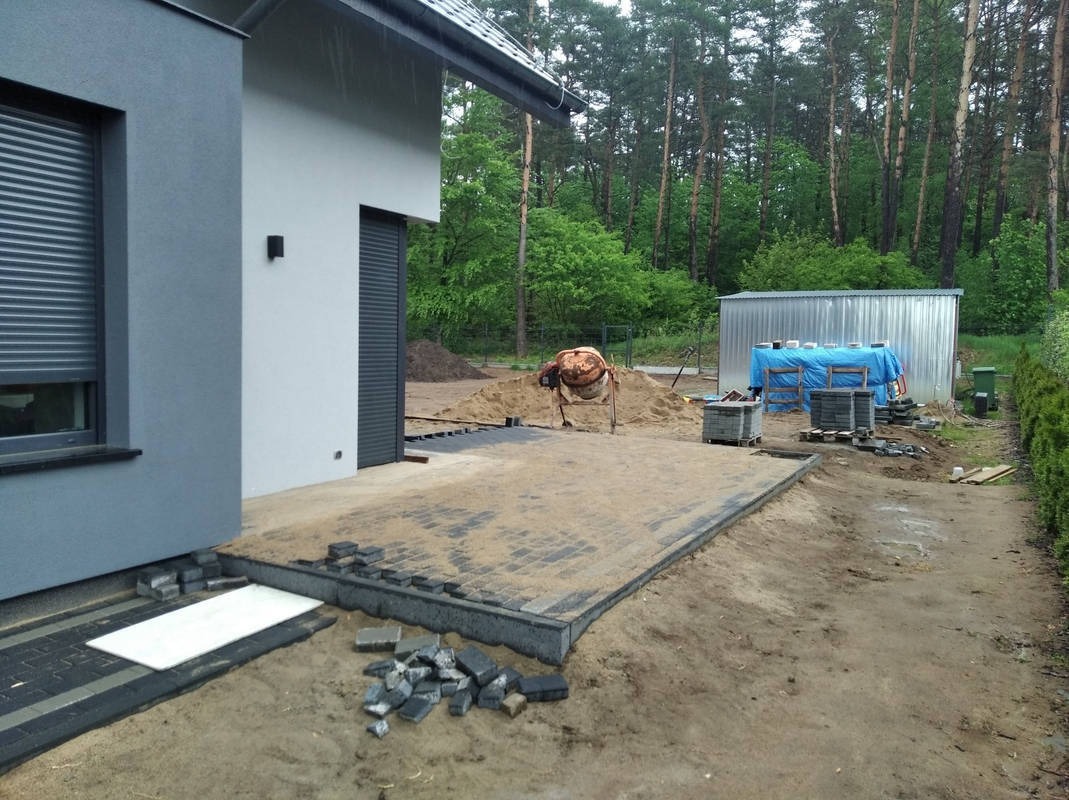 Układanie kostki brukowej w trakcie prac: widoczny fragment ułożonej kostki, podsypka piaskowa, obrzeża oraz sterta kostki brukowej obok budynku z szarą elewacją i roletami zewnętrznymi, betoniarka...