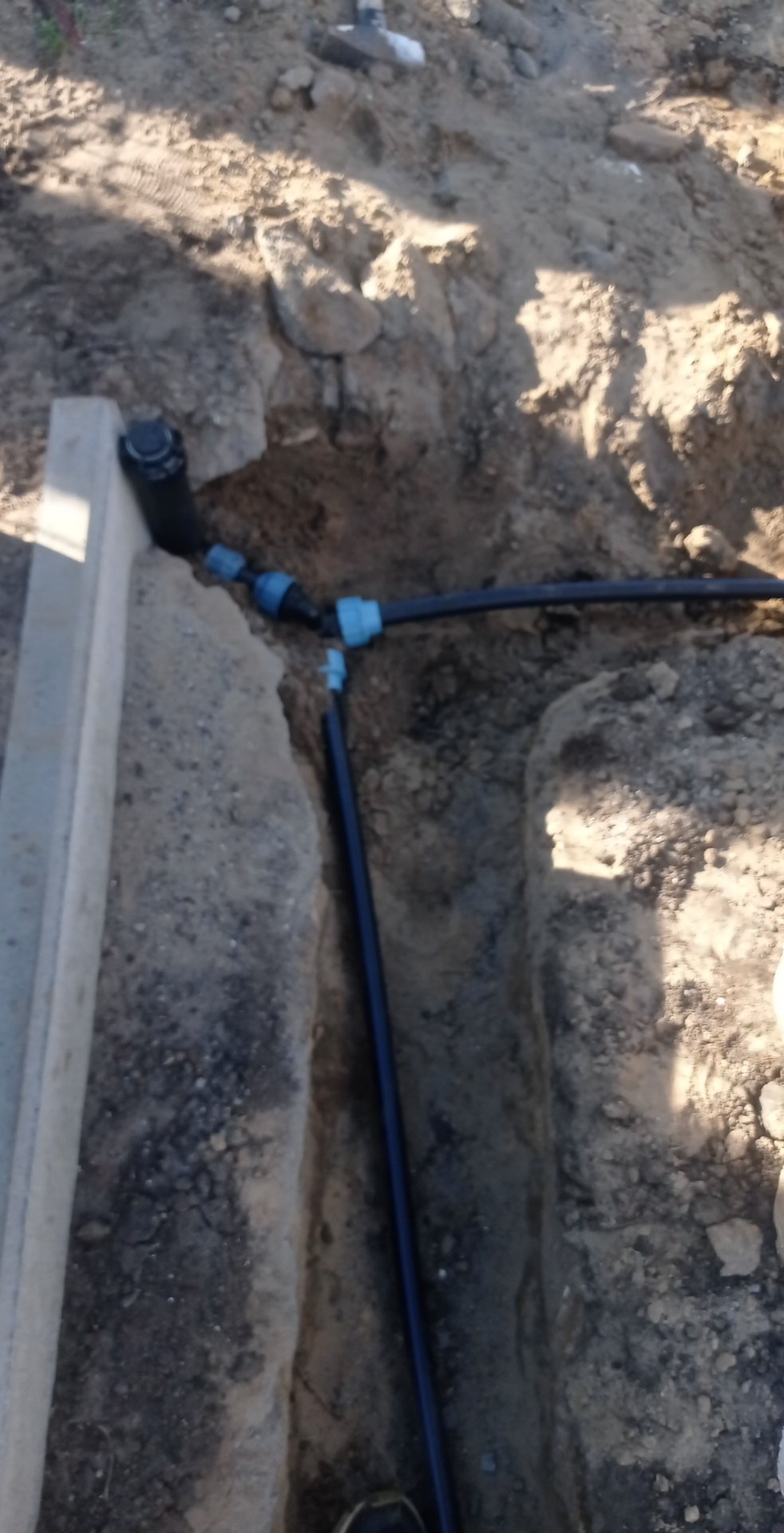 Wykop w ziemi z ułożonymi rurami z niebieskimi złączkami, widoczny betonowy obrzeże.