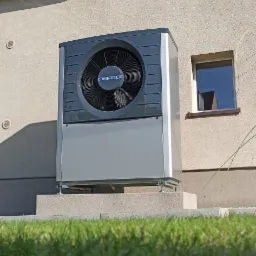 Zewnętrzna, szara pompa ciepła marki DEFRO zamontowana na betonowym podwyższeniu przy ścianie budynku z oknem, widok z poziomu trawy.