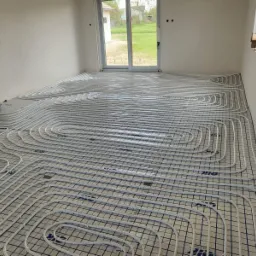 Białe rury grzewcze ułożone spiralnie na niebieskiej macie w surowym pomieszczeniu z widokiem na zewnątrz przez okno balkonowe.