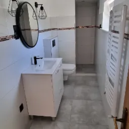 Nowoczesna łazienka z szarymi płytkami podłogowymi i białymi płytkami ściennymi z brązowym paskiem dekoracyjnym, umywalką z czarną baterią, okrągłym lustrem z czarną ramą, czarnymi lampami...