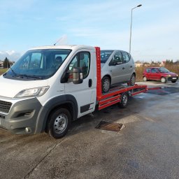 Hsm Marcin Ignasiak - Biała laweta z załadowanym srebrnym samochodem osobowym, na parkingu w tle widoczny czerwony samochód osobowy.