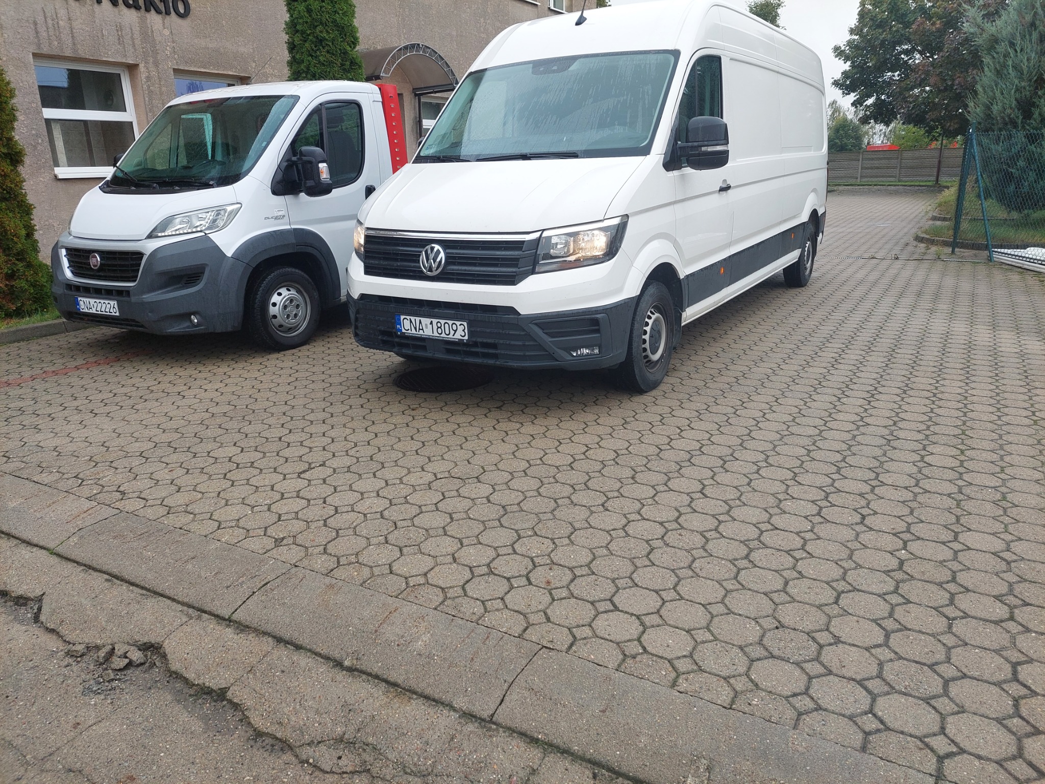 Dwa białe busy dostawcze zaparkowane na brukowanym placu przed budynkiem w pochmurny dzień.