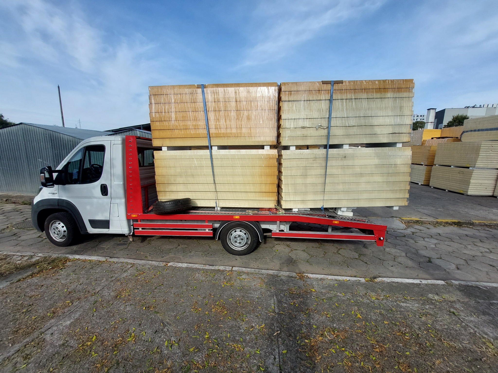 Biała laweta z czerwoną platformą przewozi zabezpieczone folią i pasami pakiety płyt izolacyjnych na tle magazynu i błękitnego nieba.