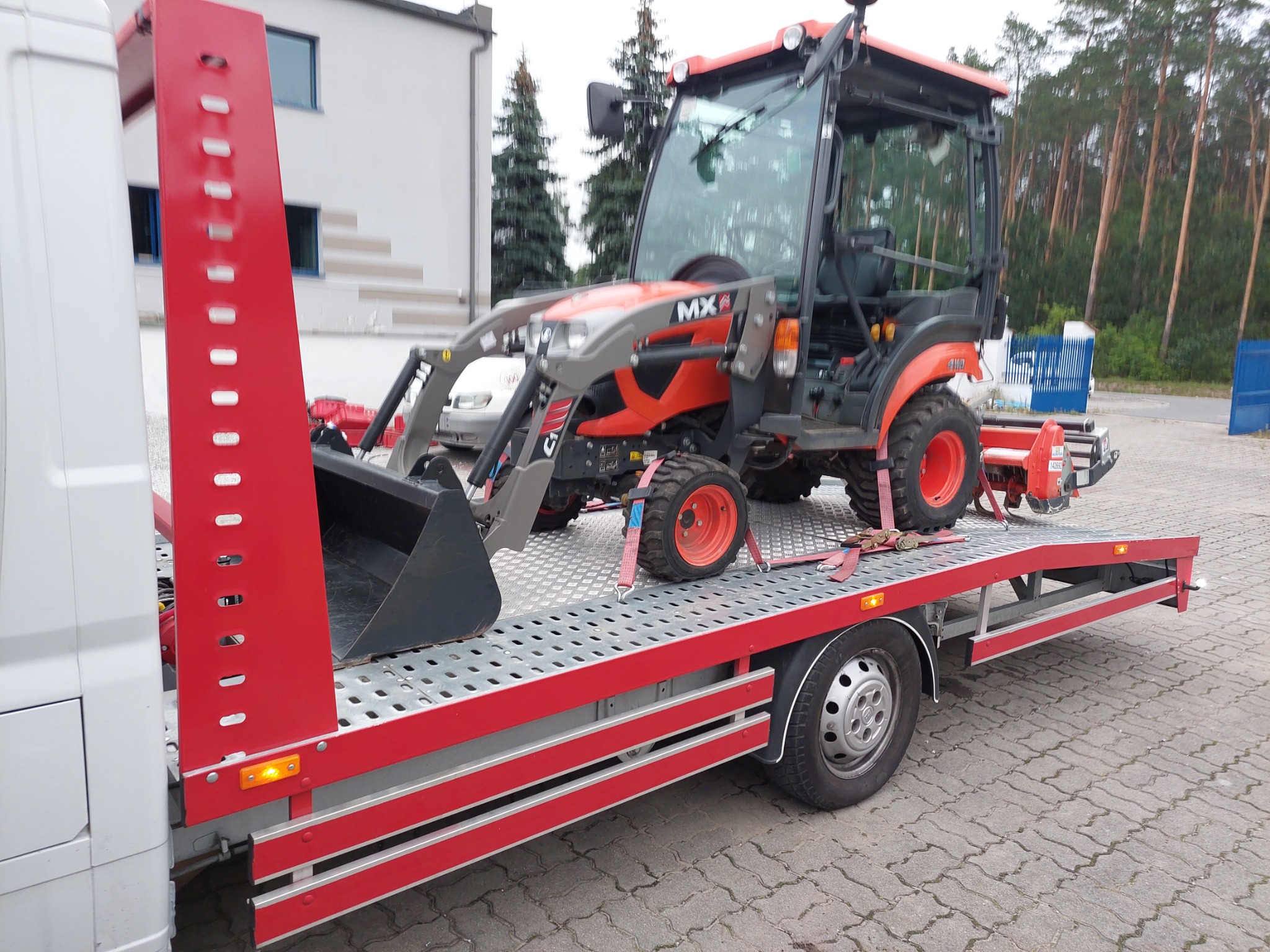 Pomarańczowo-szary traktor z łyżką załadowany na lawetę z czerwonymi elementami mocowany pasami transportowymi, na tle budynku i drzew.