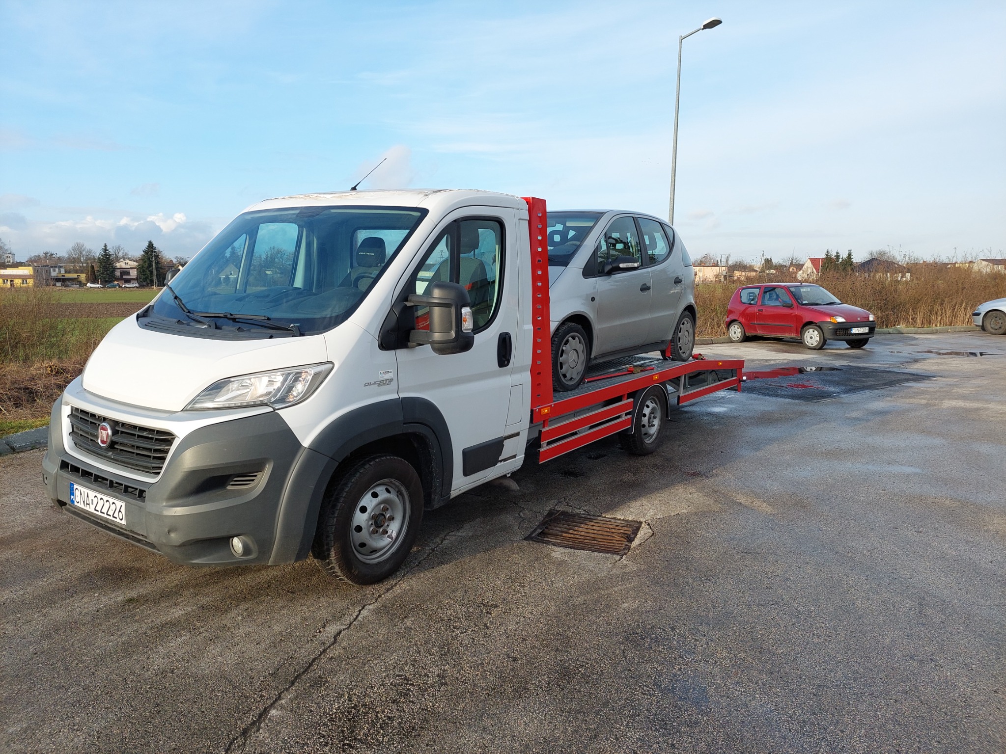 Biała laweta z załadowanym srebrnym samochodem osobowym, na parkingu w tle widoczny czerwony samochód osobowy.
