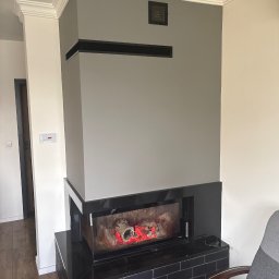 Artur Przepiórka - Szara ściana z kominkiem elektrycznym i dekoracyjną sztukaterią sufitową. Widoczny fragment fotela i drzwi w tle. Nowoczesny design wnętrza.