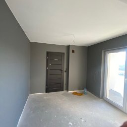 Artur Przepiórka - Pomieszczenie po malowaniu ścian na szaro, widoczne drzwi i okno balkonowe. Podłoga zabezpieczona folią malarską. Świeżo pomalowane ściany i sufit.