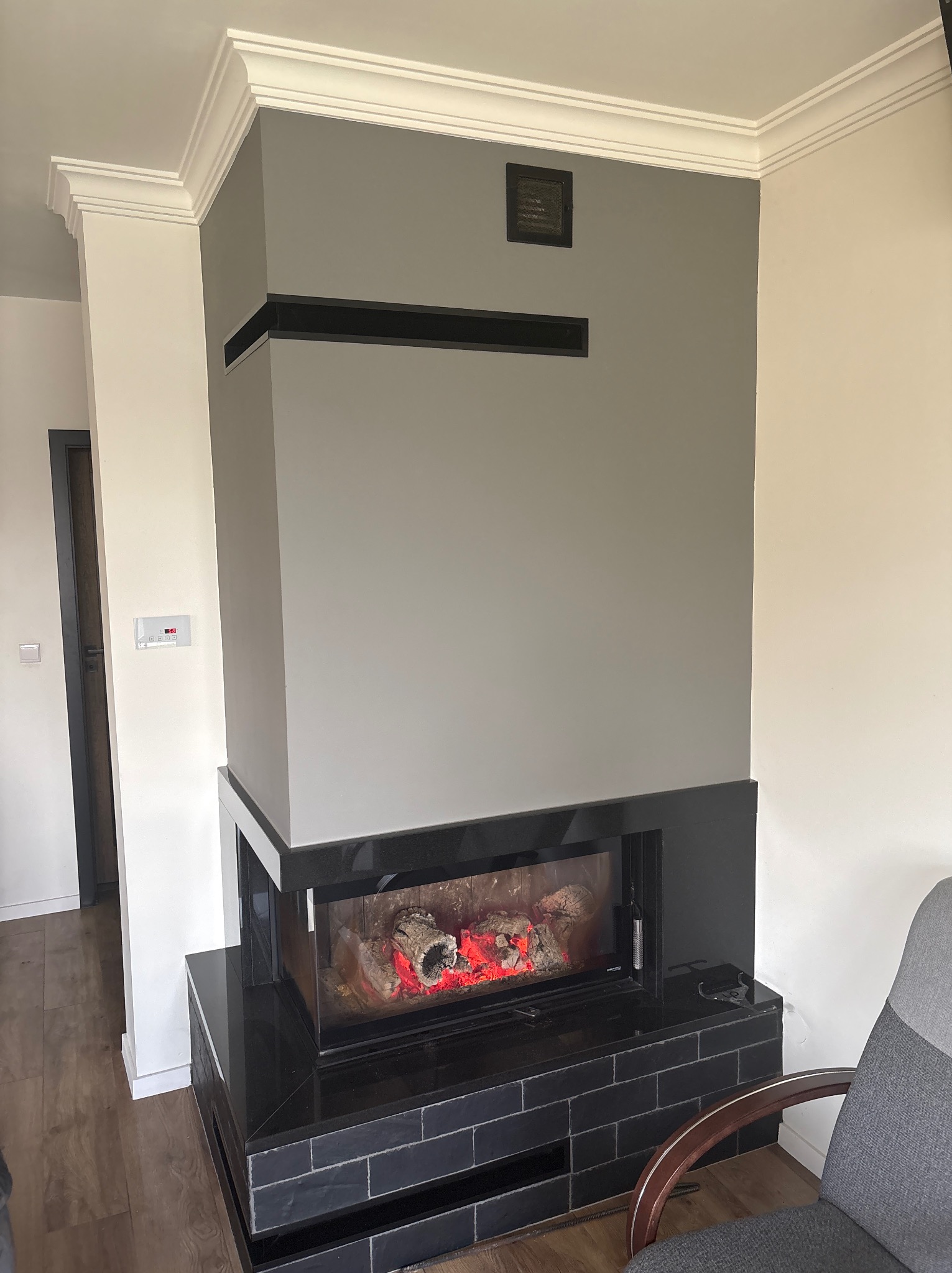 Szara ściana z kominkiem elektrycznym i dekoracyjną sztukaterią sufitową. Widoczny fragment fotela i drzwi w tle. Nowoczesny design wnętrza.