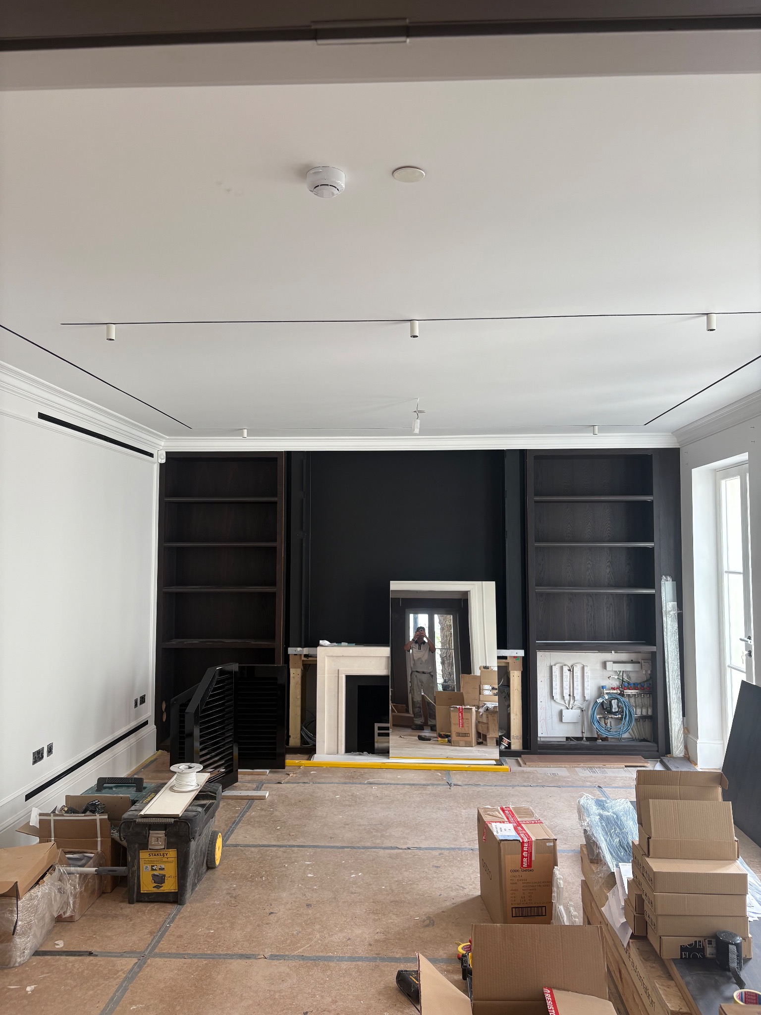 Wnętrze w trakcie remontu: widoczne regały, kominek, instalacje i narzędzia. Podłoga zabezpieczona, kartony i skrzynka narzędziowa na pierwszym planie.