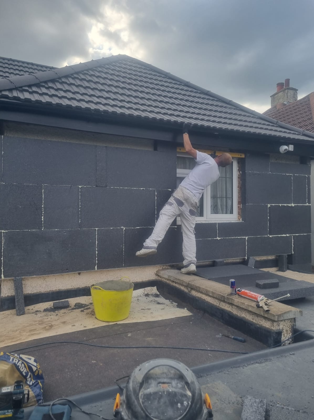 Pracownik montuje izolację termiczną na ścianie budynku. Widoczny żółty pojemnik z zaprawą, narzędzia i fragment dachu. Praca na wysokości, zabezpieczenia.