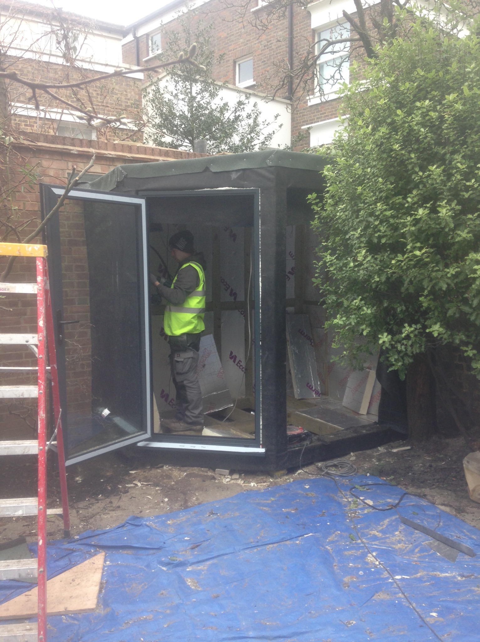 Budowa małej konstrukcji szkieletowej w ogrodzie. Pracownik w kamizelce odblaskowej montuje izolację wewnątrz. Widoczna drabina i plandeka.