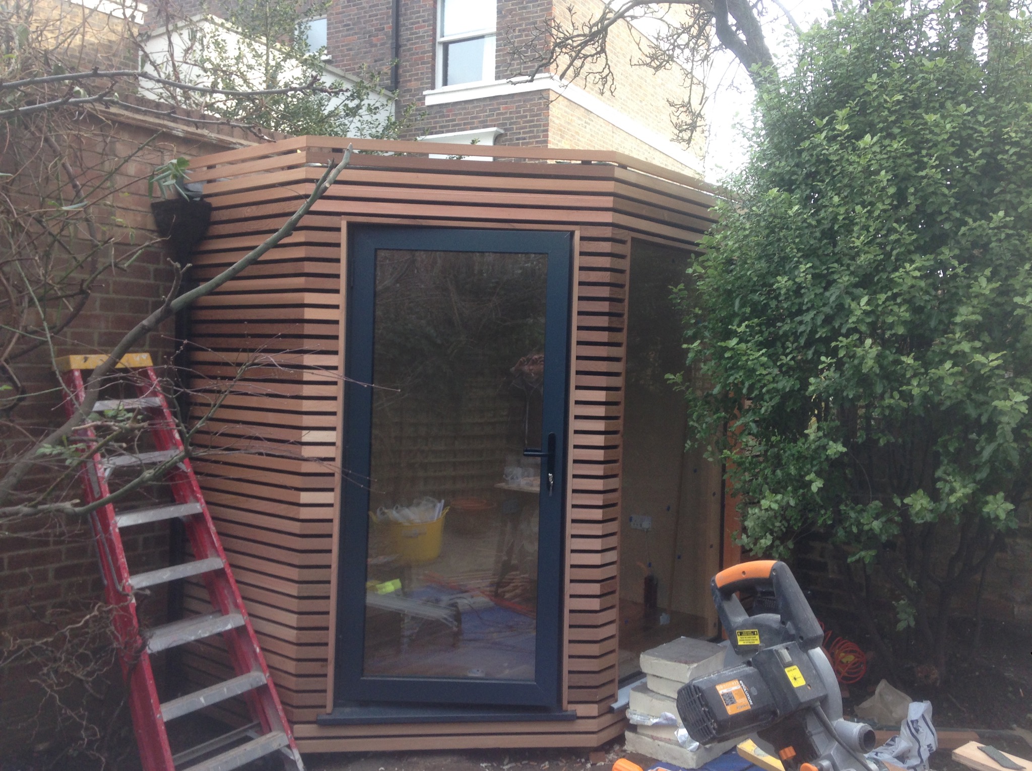 Nowoczesna altana ogrodowa z drewnianą elewacją, widoczna drabina i narzędzia, sugerujące etap budowy lub wykończenia. Ujęcie z zewnątrz, w tle budynek mieszkalny.