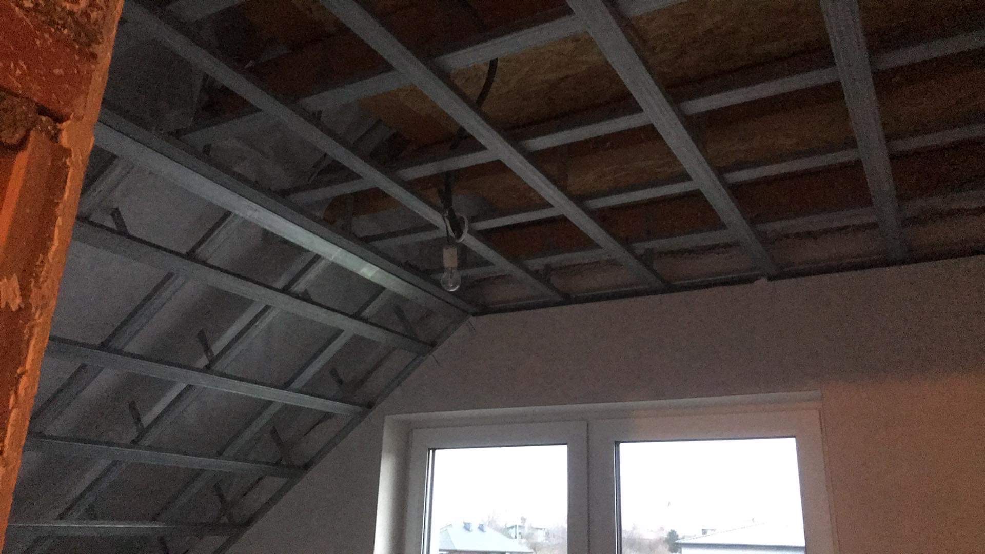 Sufit podwieszany z metalową konstrukcją w trakcie montażu, widok z dołu, surowy stan, okno w tle.