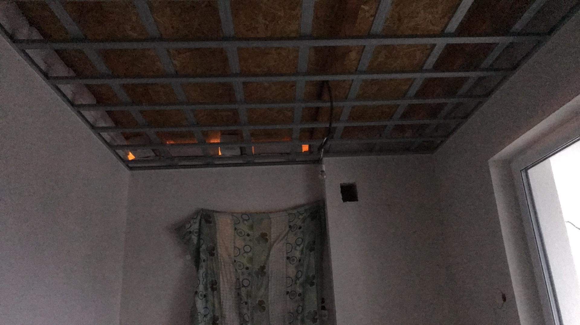 Montaż sufitu podwieszanego z widoczną konstrukcją metalową i izolacją, w tle fragment okna i ściany z zasłoniętym otworem.