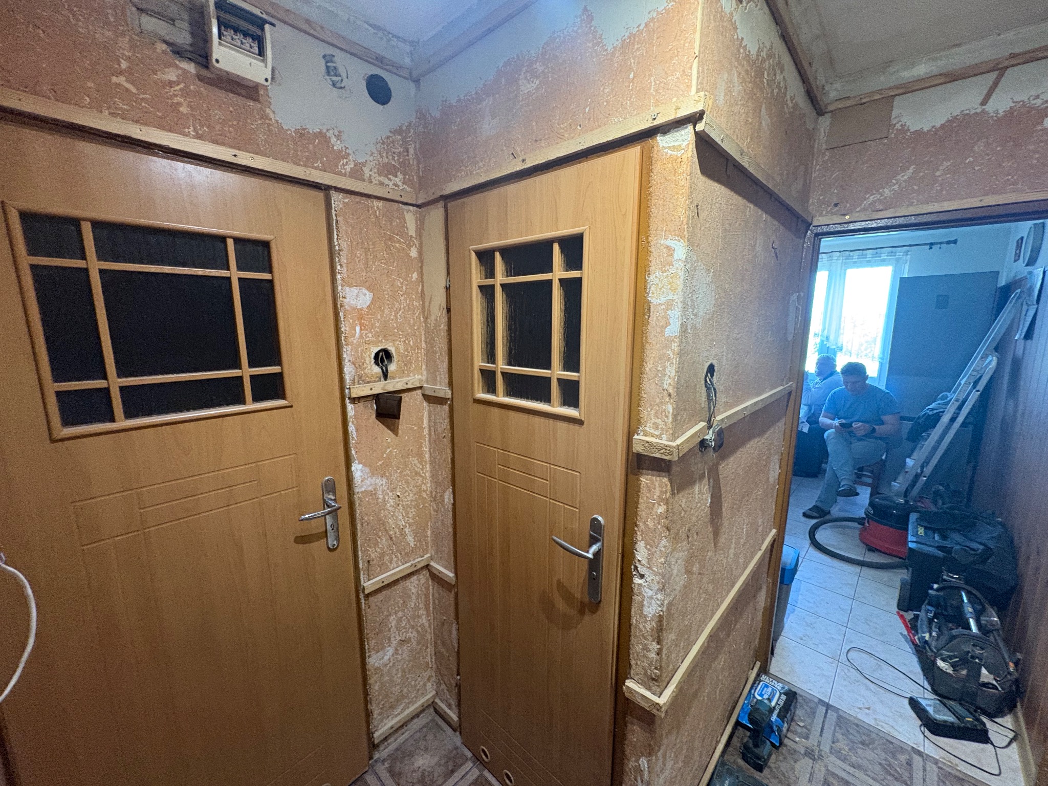 Korytarz w trakcie remontu: ściany ze śladami starej farby, dwie drewniane drzwi z szybkami, widoczne narzędzia i osoba w tle. Przygotowanie do prac wykończeniowych.