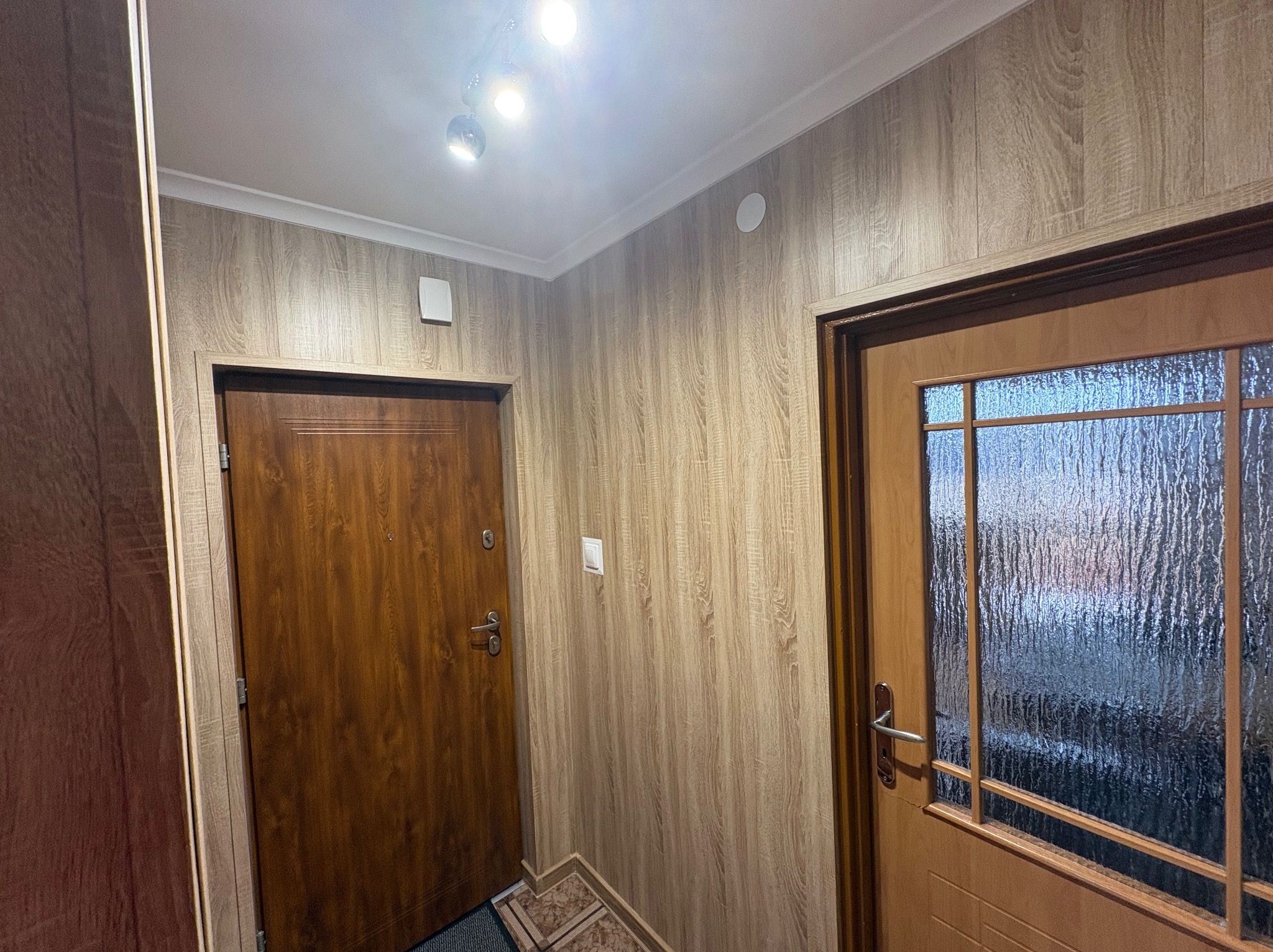 Korytarz po remoncie: ściany wyłożone panelami imitującymi drewno, dwoje drewnianych drzwi, oświetlenie sufitowe. Widoczne detale wykończenia.