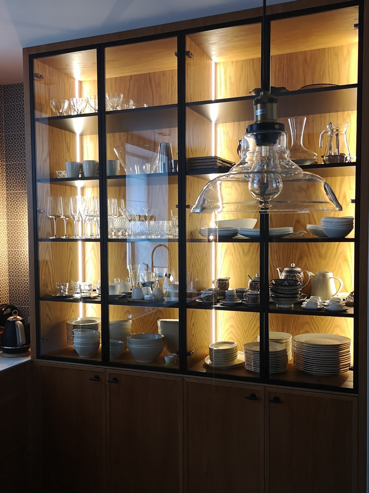 Wysoka, przeszklona witryna z czarnymi ramami i drewnianymi plecami, oświetlona pionowymi paskami LED, wypełniona zastawą stołową, szkłem i porcelaną, z wiszącą lampą w stylu industrialnym.
