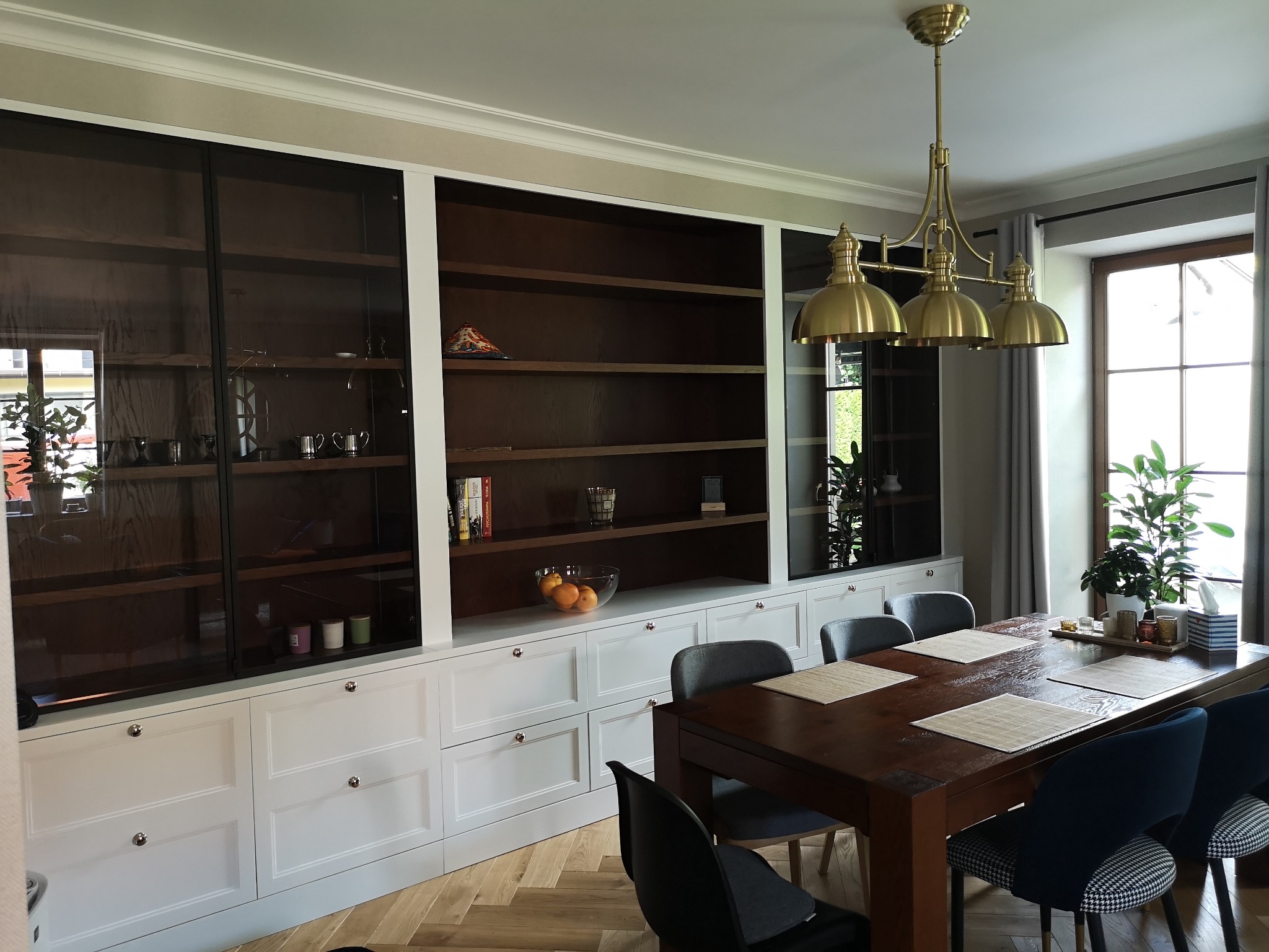 Jadalnia z zabudową meblową w stylu klasycznym: białe szafki z szufladami i przeszklone witryny, drewniany stół z nakryciami i krzesłami, złota lampa wisząca.