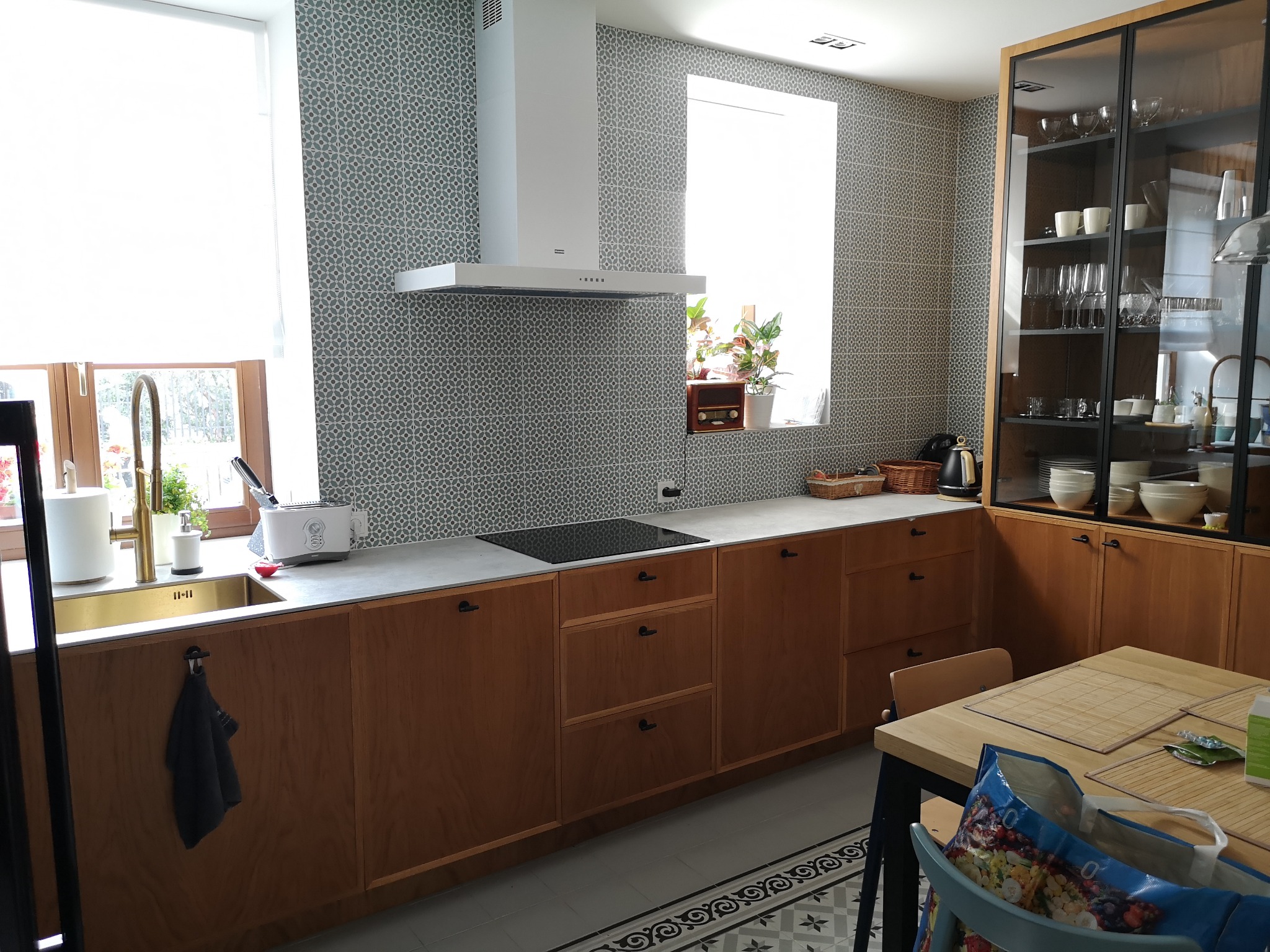 Nowoczesna kuchnia z drewnianymi frontami, szarym blatem i wzorzystą ścianą, wyposażona w złoty zlew i biały okap.