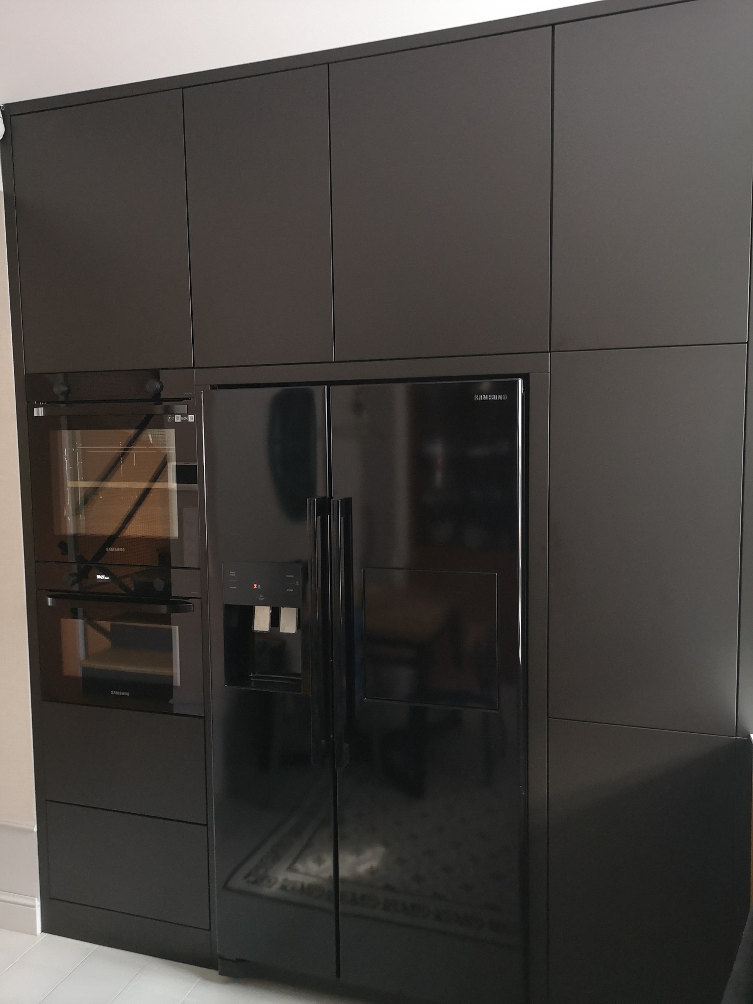 Ciemna zabudowa kuchenna z piekarnikiem i lodówką side-by-side, z minimalistycznymi frontami bez uchwytów, odbijająca światło w połyskującej powierzchni.