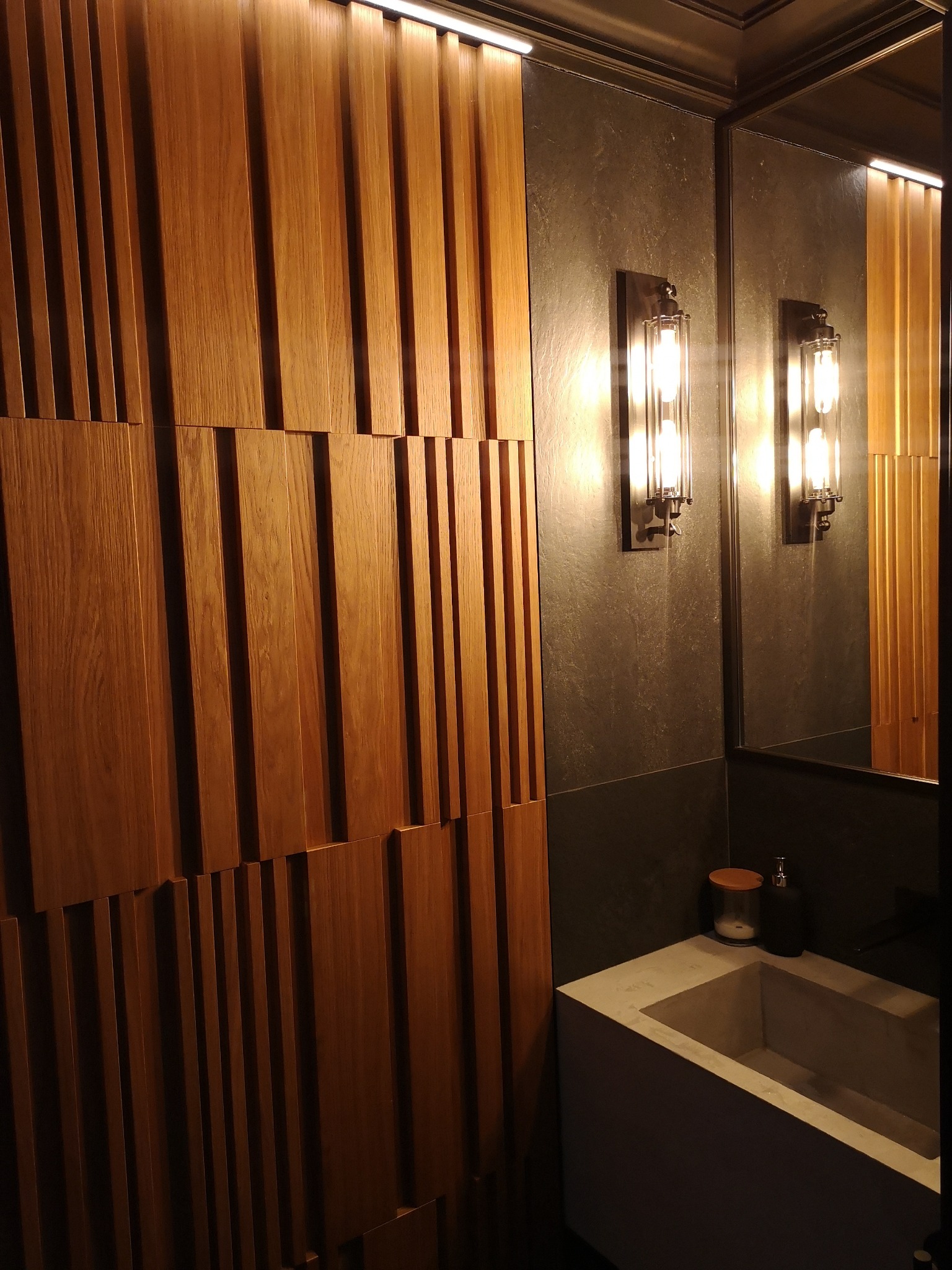Nowoczesna łazienka z drewnianą ścianą z pionowych paneli, szarą płytką, lustrem i designerskimi lampami ściennymi.