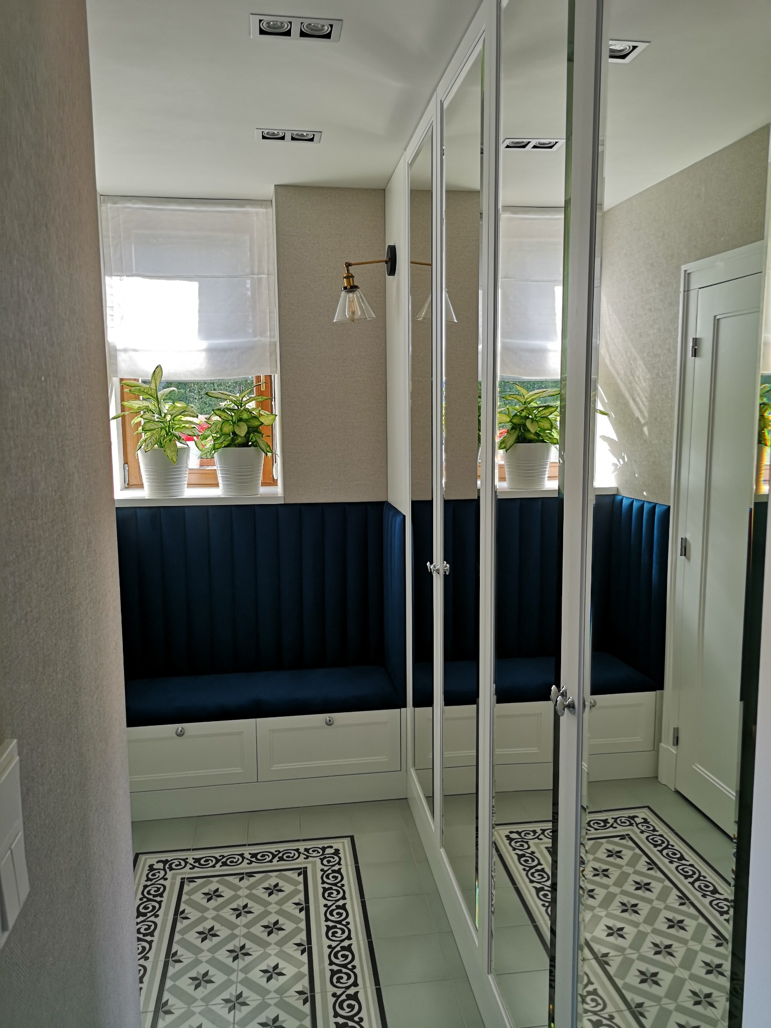 Wnętrze przedpokoju z zabudową wnękową: siedzisko z granatową tapicerką pod oknem, szafa z lustrzanymi drzwiami, wzorzysta podłoga.