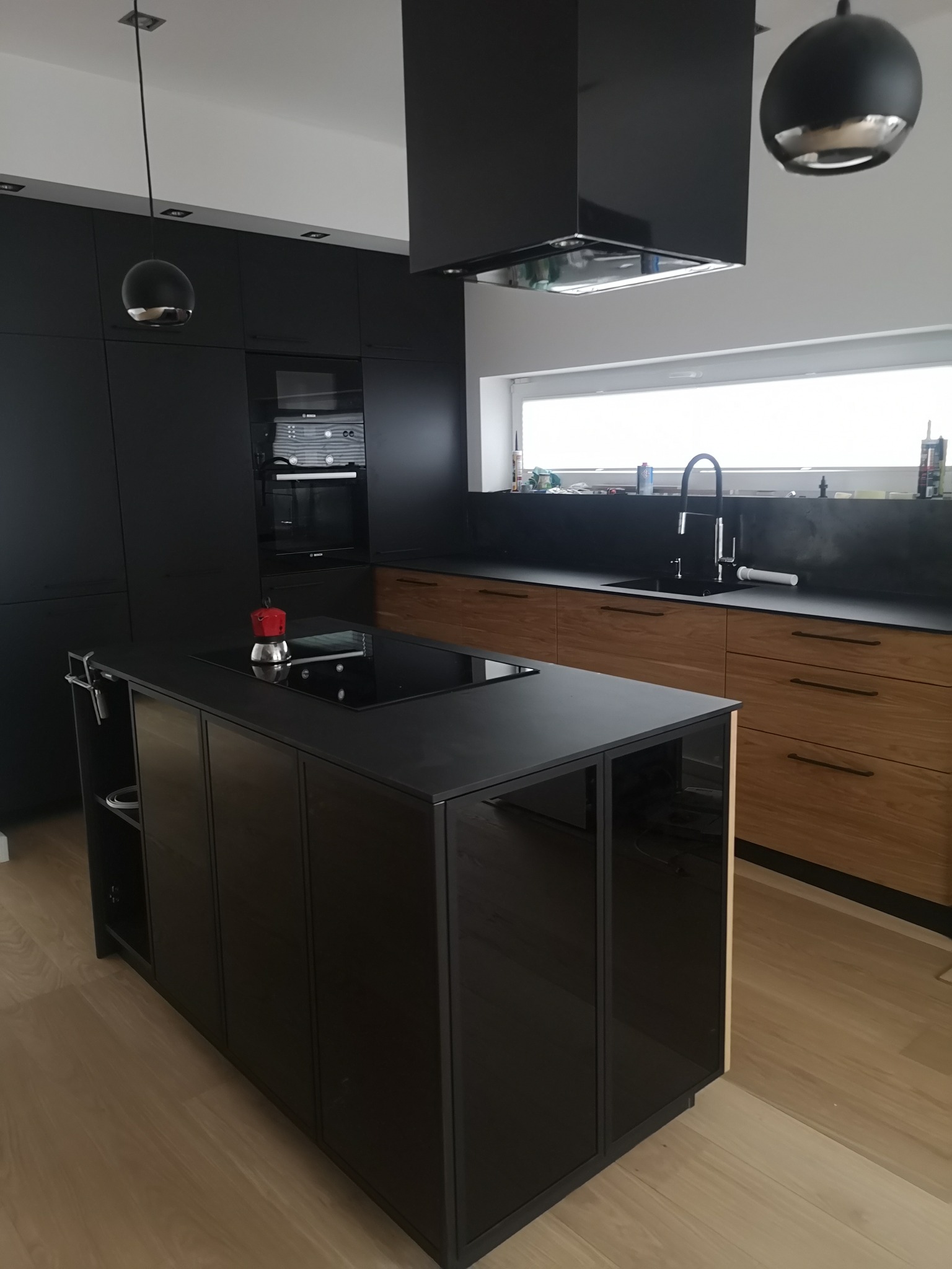 Nowoczesna kuchnia z czarnymi frontami i drewnianymi szufladami, wyspa kuchenna z płytą indukcyjną i czerwonym ekspresem do kawy, minimalistyczne oświetlenie.