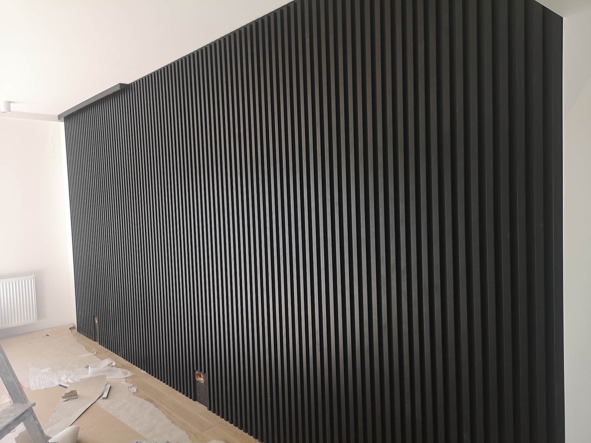 Ściana wykończona czarnymi, pionowymi lamelami drewnianymi, widoczne niedokończone prace wykończeniowe na podłodze.