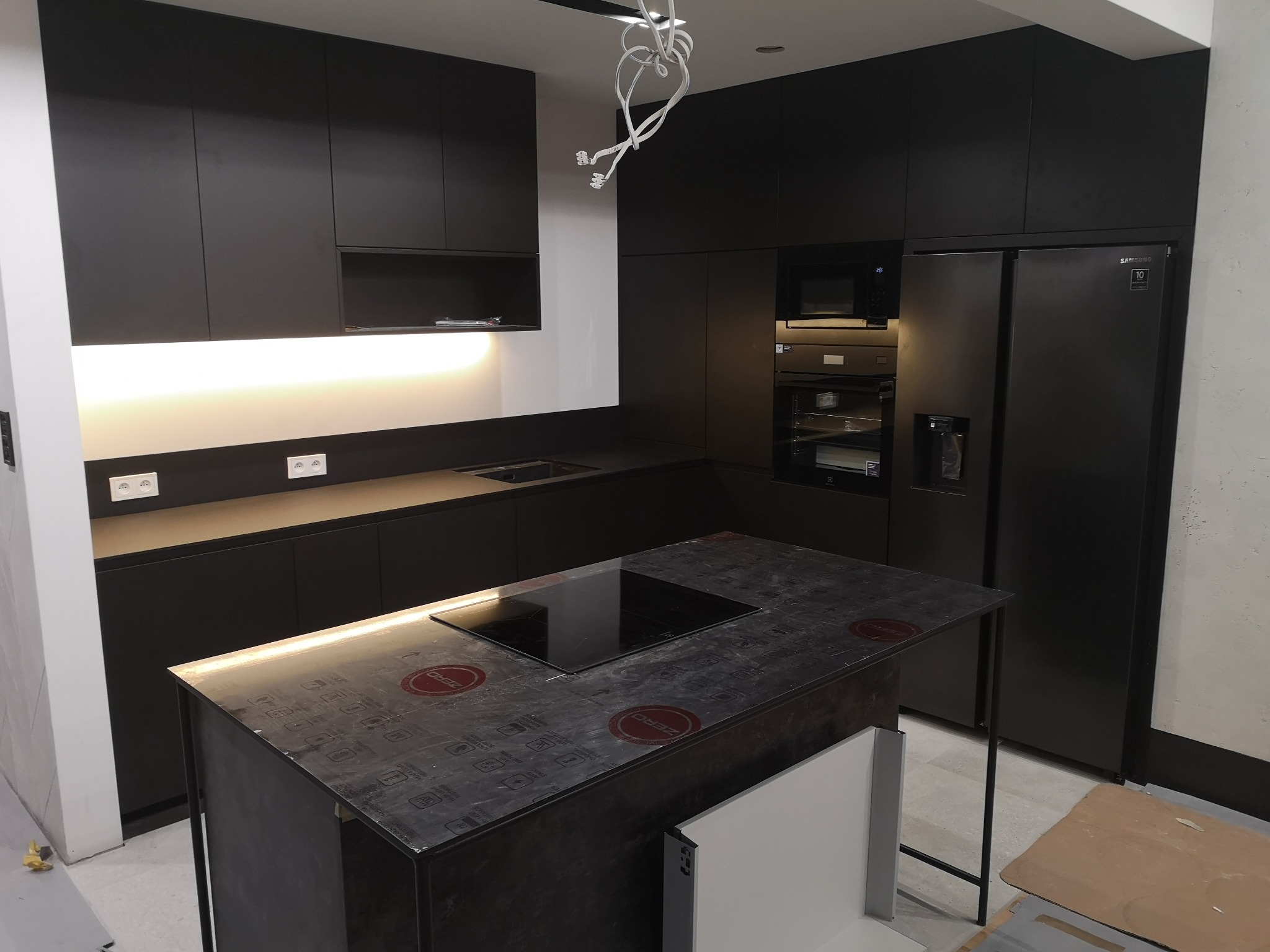 Nowoczesna, czarna kuchnia z wyspą i oświetleniem LED pod szafkami, płyta indukcyjna na wyspie zabezpieczona folią. W tle zabudowa kuchenna z piekarnikiem i lodówką.