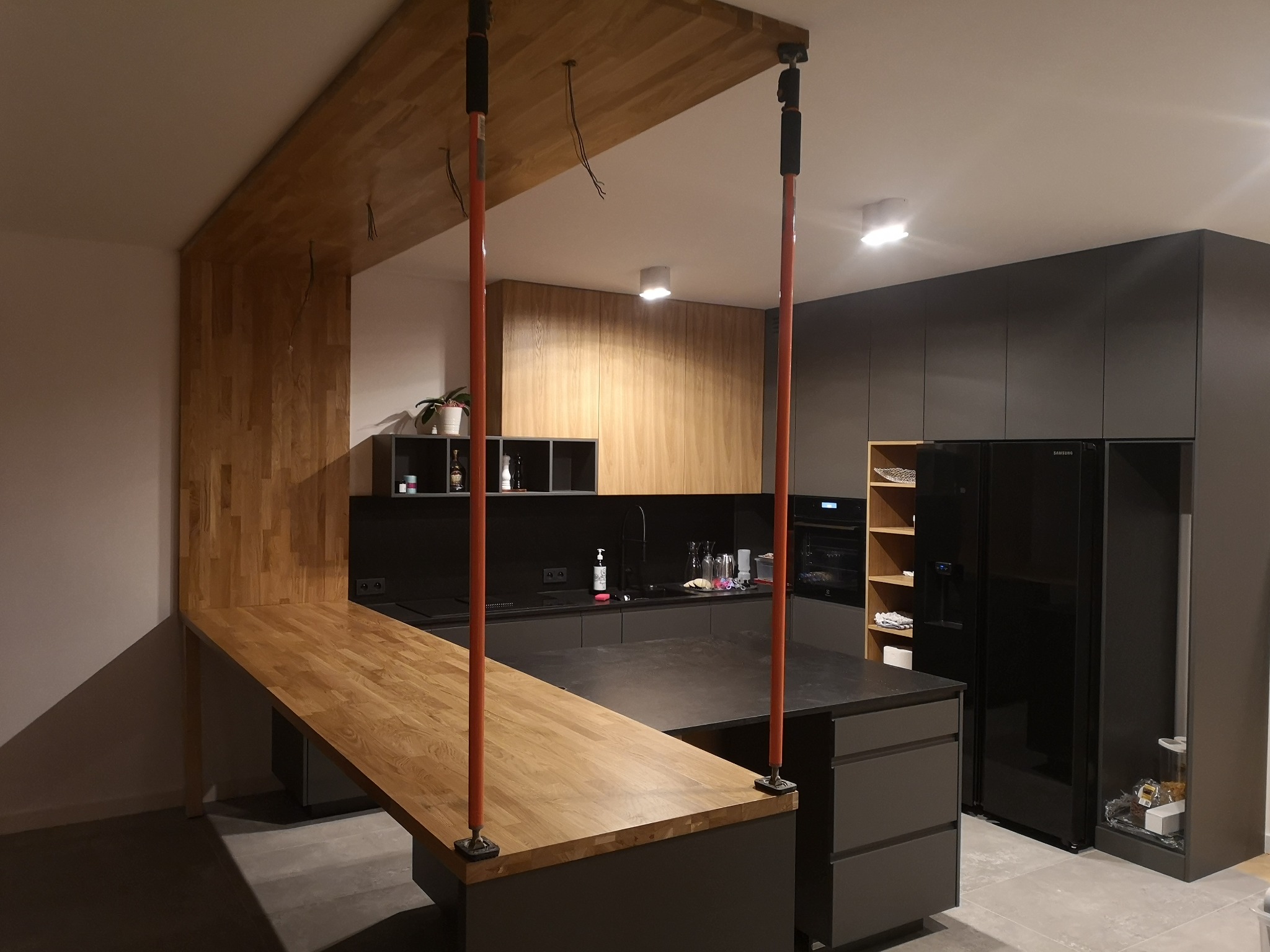Nowoczesna kuchnia z drewnianym blatem przechodzącym w ściankę działową, oświetlenie punktowe, ciemne szafki i sprzęt AGD, widoczne przewody elektryczne z sufitu.
