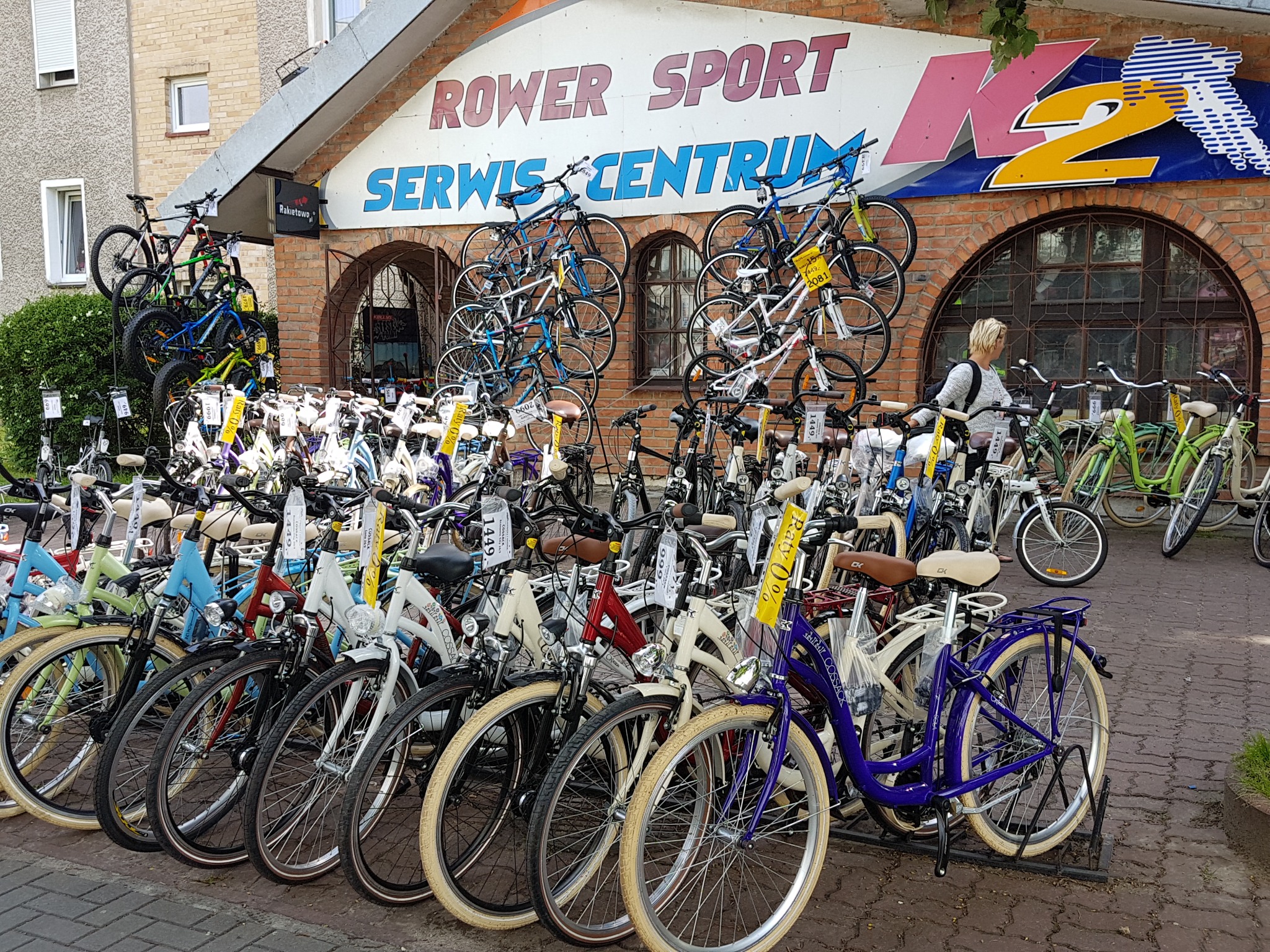 Wystawa rowerów różnego typu i koloru przed sklepem Rower Sport Serwis Centrum, z widocznym logo K2 na budynku i klientką przeglądającą asortyment.