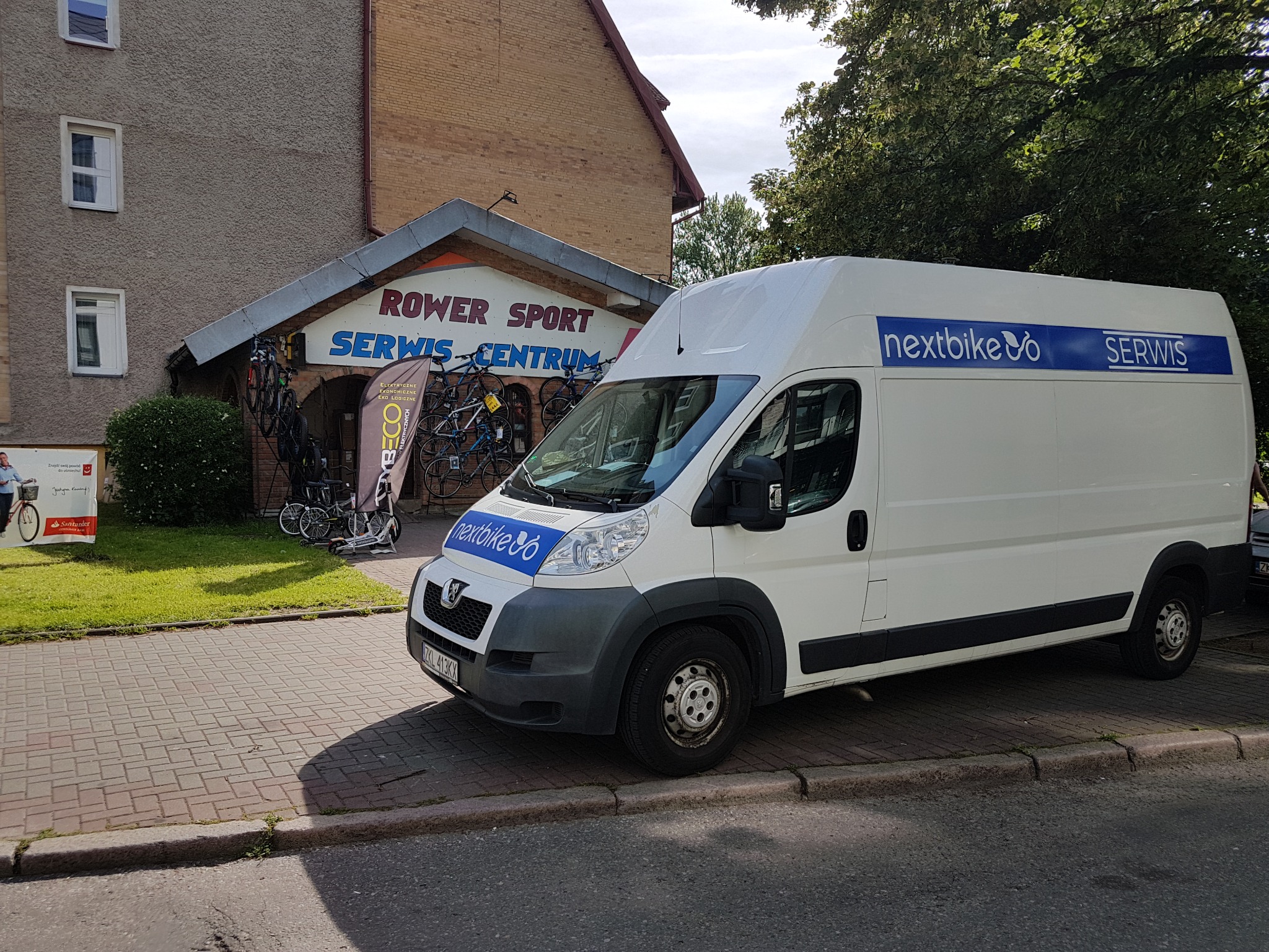 Samochód dostawczy firmy Nextbike z Kołobrzegu zaparkowany przed serwisem rowerowym Rower Sport Serwis Centrum. Na dachu i boku pojazdu widoczne logo i napis SERWIS.