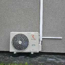 Biały klimatyzator Rotenso IMOTO Series zamontowany na zewnątrz budynku z szarą elewacją, widoczne przewody i fragment trawnika.