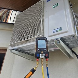 Cleanline Energy Sp. z o.o. - Biała klimatyzacja Panasonic z podłączonym manometrem cyfrowym Testo 550s na elewacji budynku. Widoczne przewody i elementy instalacji.