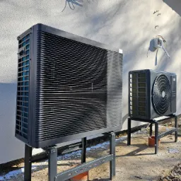 Dwie jednostki zewnętrzne systemu grzewczego Panasonic, ustawione na metalowych wspornikach przed białą ścianą budynku z widoczną puszką elektryczną, w trakcie lub tuż po instalacji.