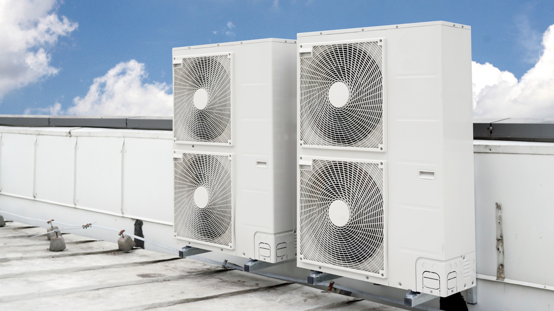 Dwa białe, bliźniacze klimatyzatory przemysłowe zamontowane na płaskim dachu z widocznymi elementami mocującymi i niebem w tle.