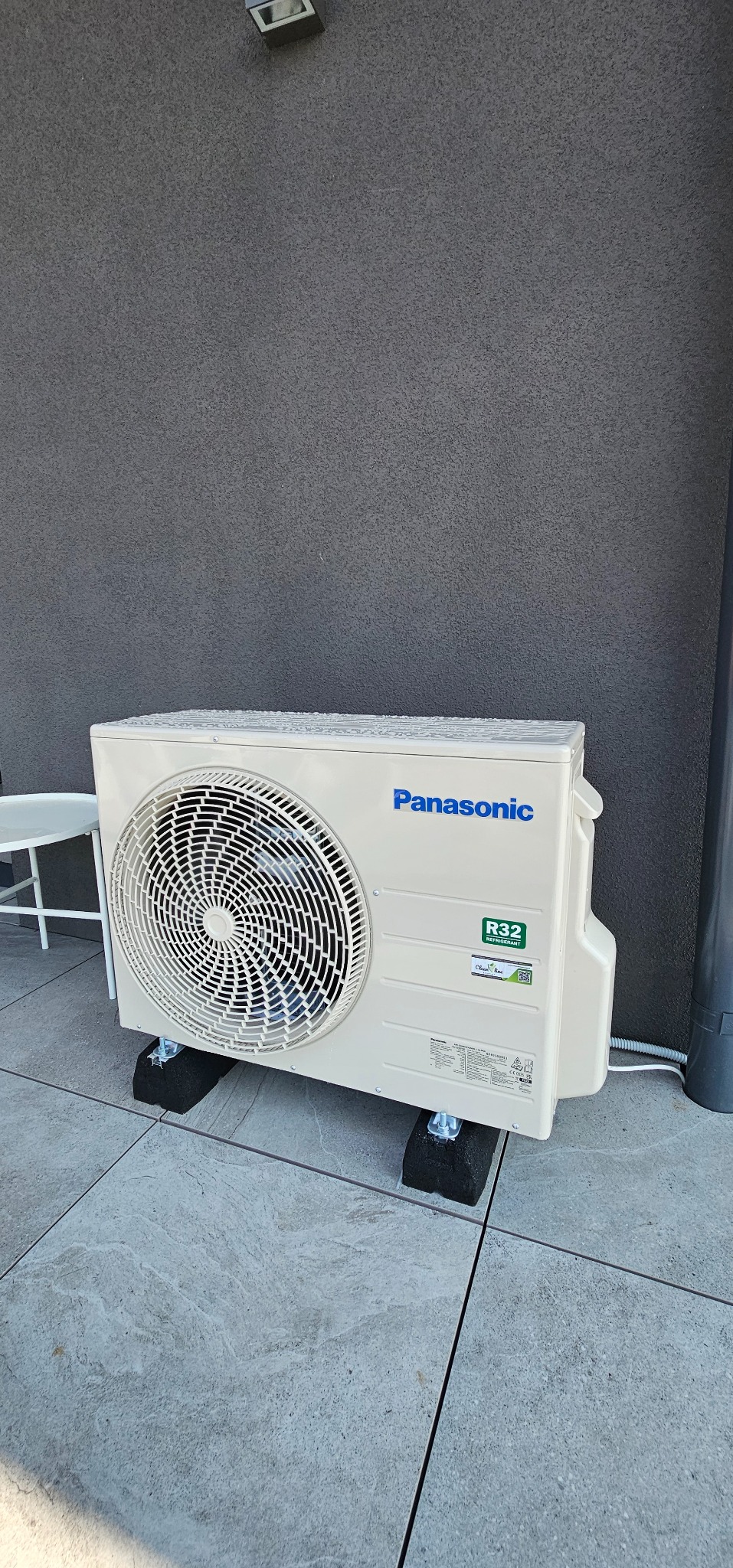 Biała jednostka zewnętrzna klimatyzacji Panasonic, zamontowana na czarnych podkładkach na szarym tarasie, z szarą ścianą w tle. Widoczny fragment białego krzesła.