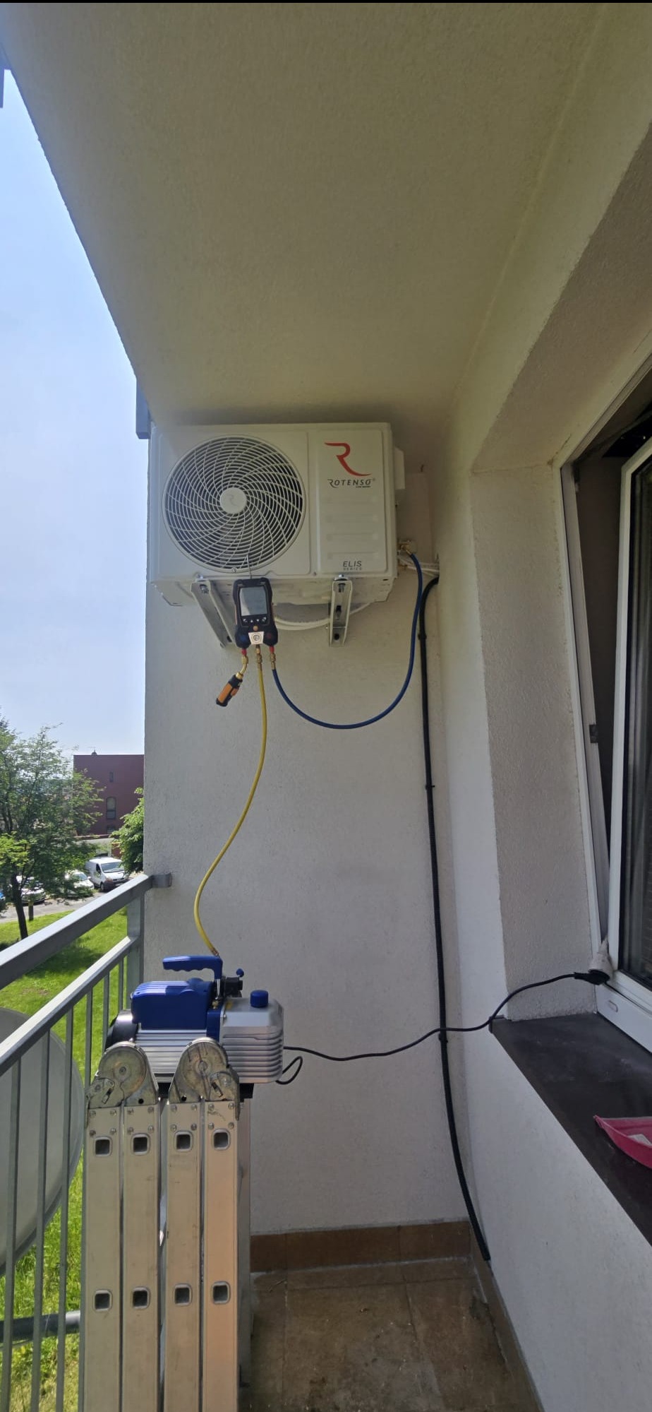 Montaż klimatyzacji Rotenso Elis na balkonie. Widoczna pompa próżniowa i manometry, co sugeruje etap serwisu lub instalacji. W tle zieleń i zabudowa miejska.