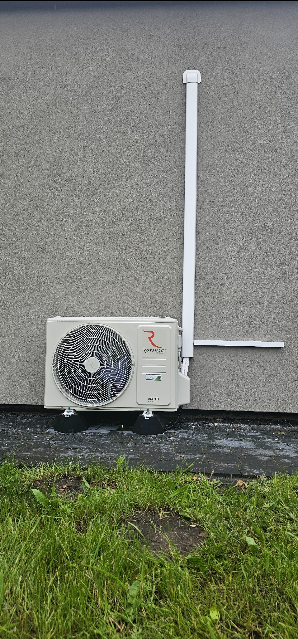 Biały klimatyzator Rotenso IMOTO Series zamontowany na zewnątrz budynku z szarą elewacją, widoczne przewody i fragment trawnika.