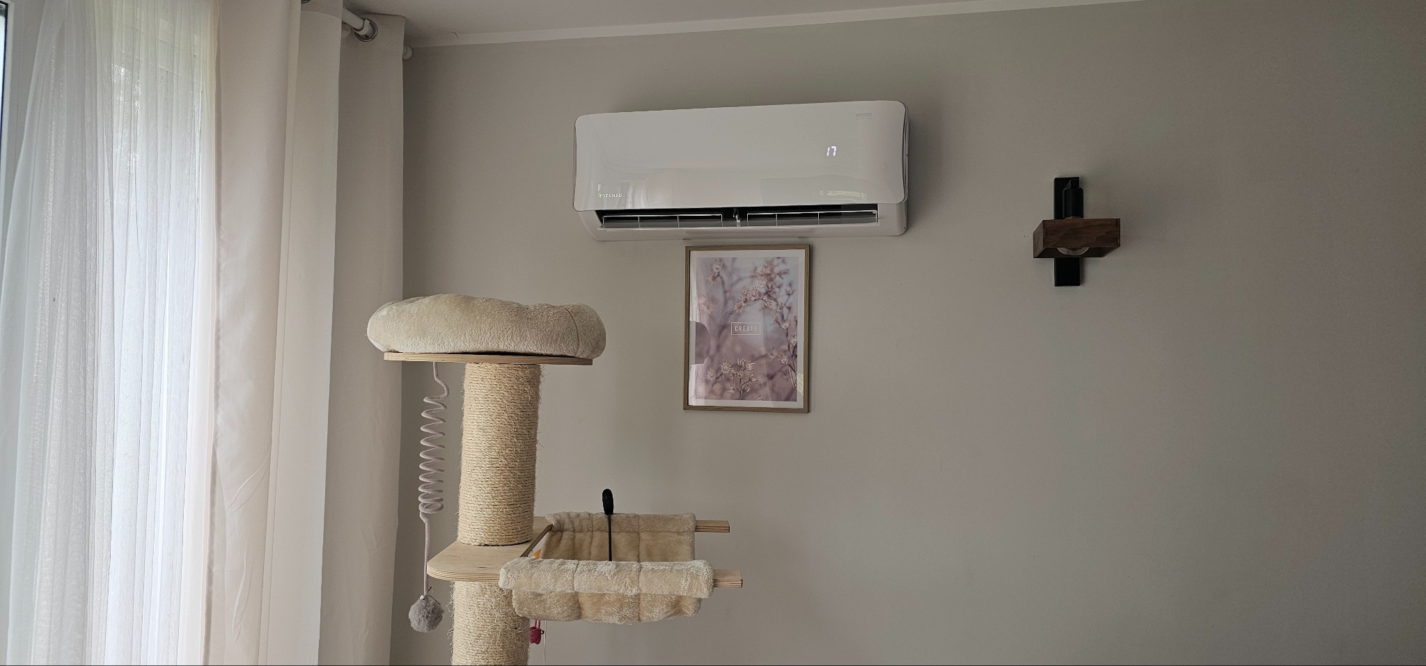 Biała klimatyzacja ścienna z wyświetlaczem temperatury, zamontowana w pokoju z drapakiem dla kota i obrazem w ramie.