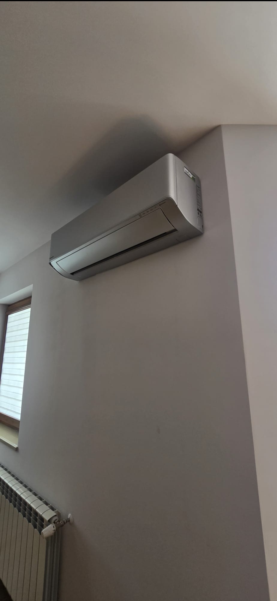 Szary klimatyzator ścienny w jasnym wnętrzu, widoczny grzejnik pod oknem. Minimalistyczny design, stonowana kolorystyka.