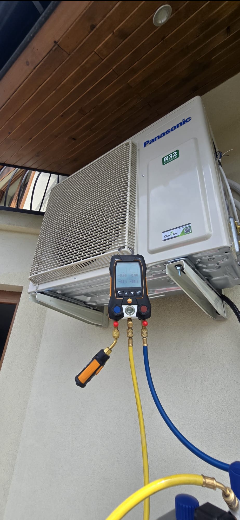 Biała klimatyzacja Panasonic z podłączonym manometrem cyfrowym Testo 550s na elewacji budynku. Widoczne przewody i elementy instalacji.