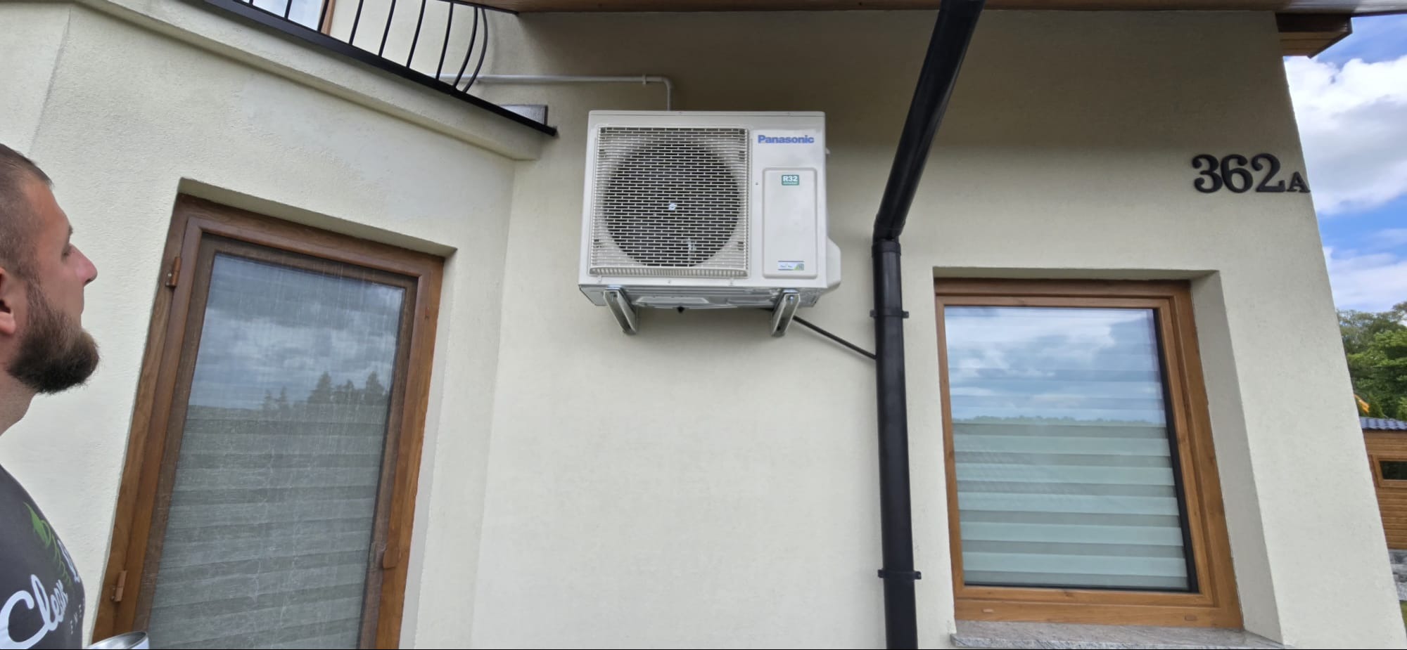 Biała klimatyzacja Panasonic zamontowana na elewacji domu, widoczny numer budynku 362a, obok okno w brązowej ramie i czarna rynna.