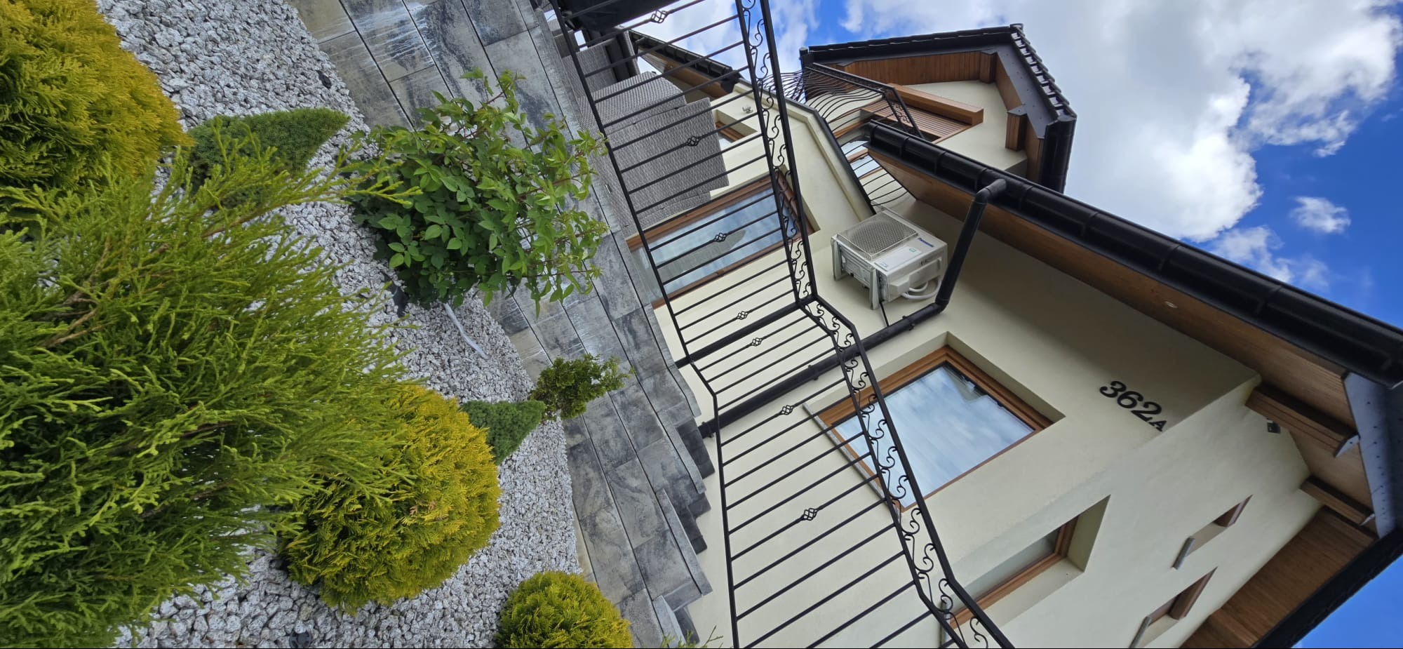 Biała jednostka klimatyzacji na elewacji domu z numerem 362A, widoczna z perspektywy ogrodu z dekoracyjnymi krzewami i kamieniami. Błękitne niebo w tle.