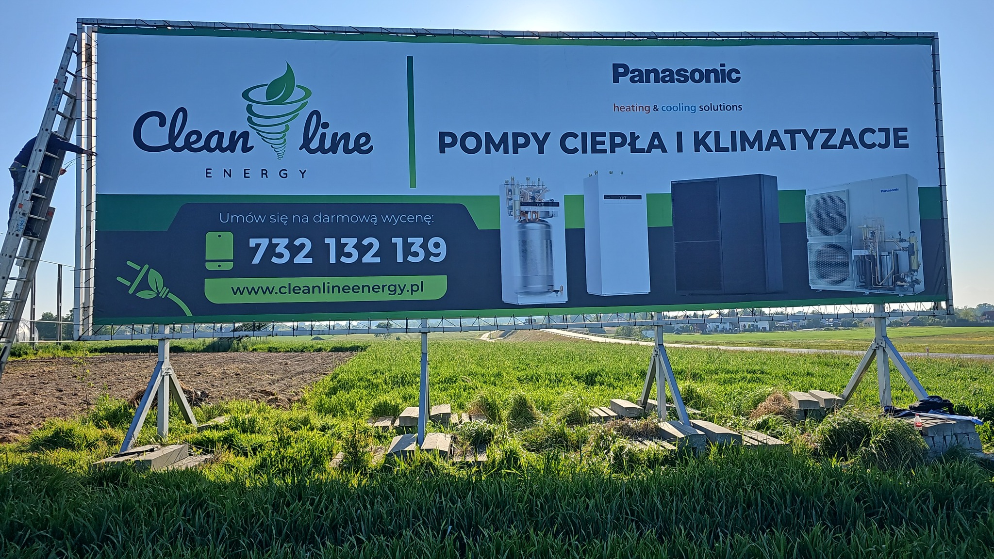 Bilbord reklamowy firmy z Ćwiklic promującej pompy ciepła i klimatyzacje Panasonic. Widoczny pracownik na drabinie montujący baner. Numer telefonu i adres strony internetowej.