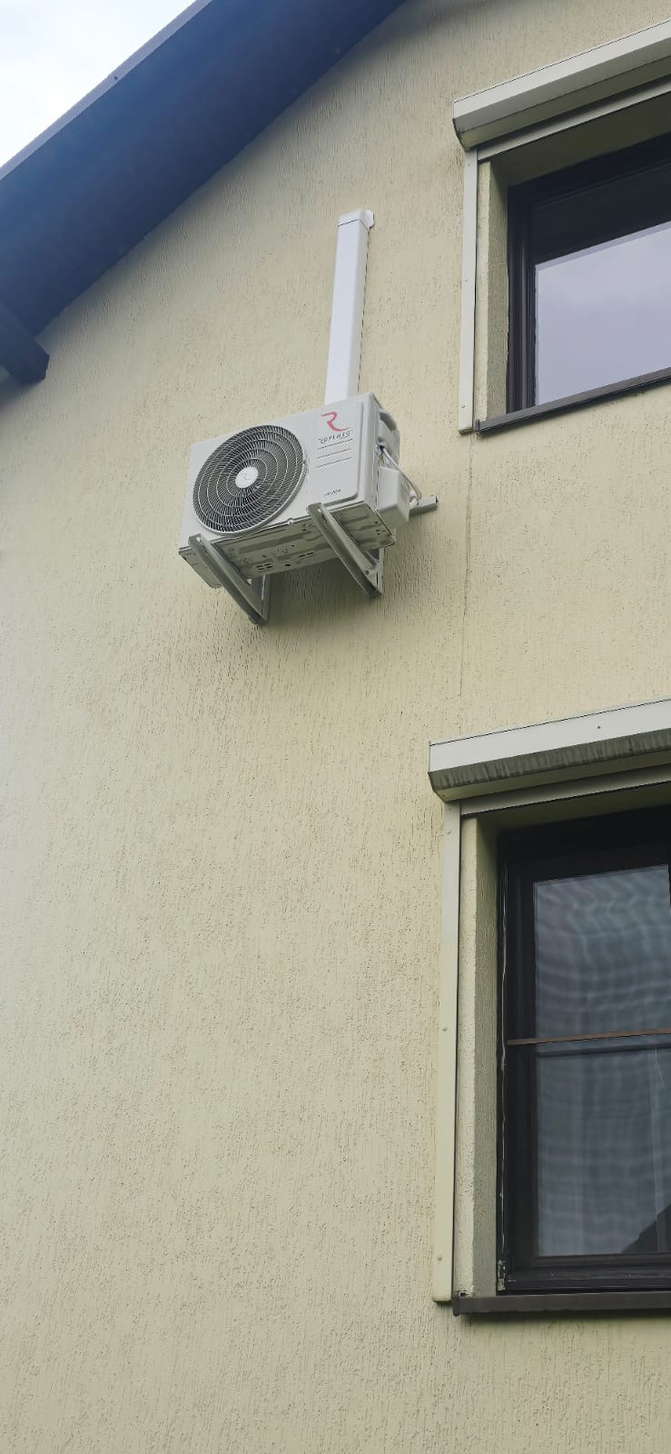 Biały klimatyzator z widocznym logo producenta, zamontowany na elewacji budynku o jasnej, chropowatej fakturze, z rurą odprowadzającą pionowo w górę.