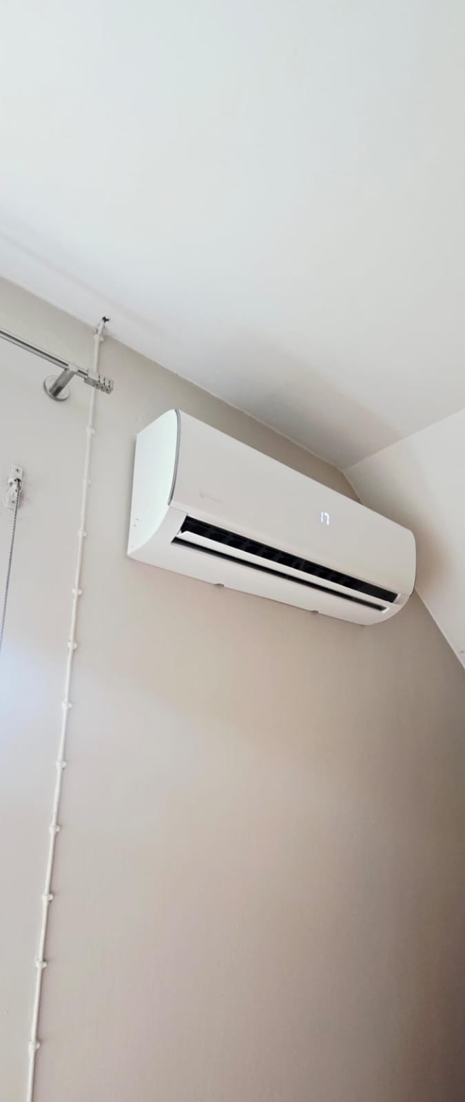 Biały klimatyzator ścienny z wyświetlaczem temperatury 17 stopni, zainstalowany w pomieszczeniu o jasnych ścianach. Widoczne przewody instalacyjne.