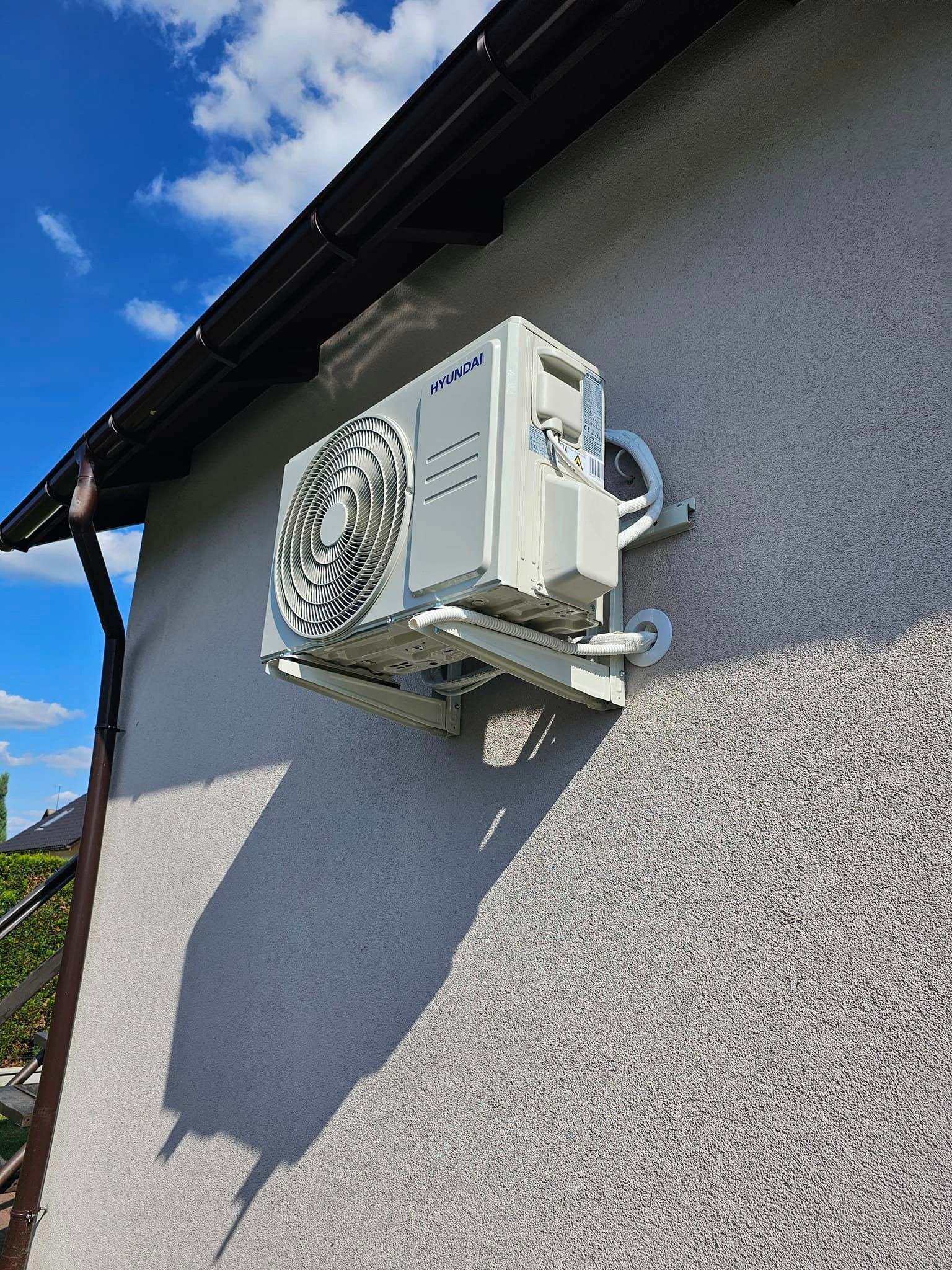 Zewnętrzna jednostka klimatyzacji Hyundai zamontowana na elewacji domu, widoczne przewody w białej izolacji i cień urządzenia na ścianie.